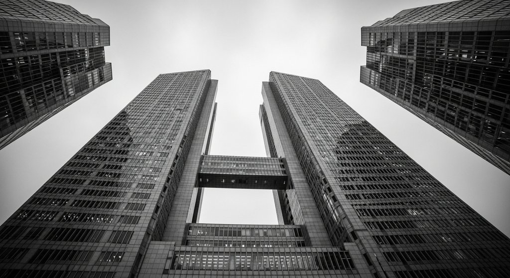 Das Gebäude der Europäischen Zentralbank in Frankfurt aus einer ungewöhnlichen Froschperspektive, aufgenommen an einem bewölkten Tag. Die Architektur wirkt imposant und modern. Das Bild ist im Stil einer analogen Schwarz-Weiß-Fotografie gehalten, mit sichtbarem Filmkorn und hohem Kontrast, was dem Motiv eine ernste und bedeutungsvolle Atmosphäre verleiht. Shot on analog film, Ilford HP5 style. →